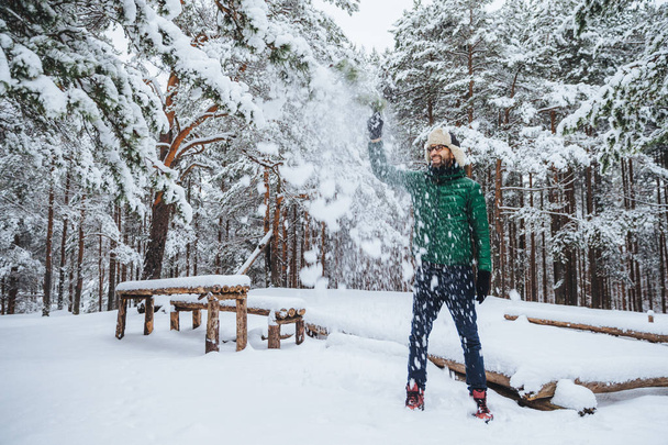 Foto al aire libre de hombre barbudo guapo vestido con ropa de abrigo, se divierte como arroja nieve en el aire, pasa las vacaciones en el bosque de invierno, expresa positividad. Concepto de recreación y clima
 - Foto, imagen
