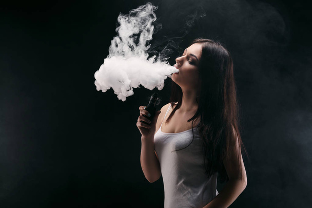 Young woman vaping e-cigarette with smoke on black - Foto, afbeelding