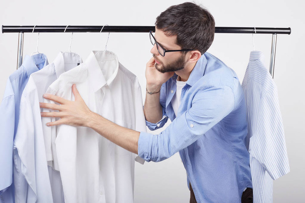 Serious homosexual male chatting over mobile phone, looking through rack with clothes, taking advice of best friend what to wear on date with his boyfriend, having special occasion. Clothing concept. - Photo, Image