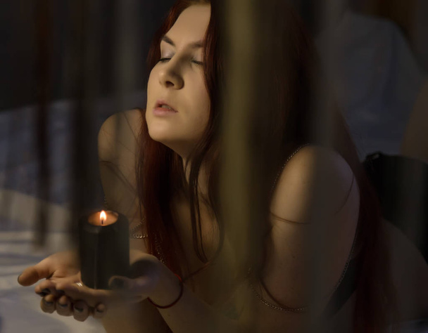 sensual redhead woman lies on a bed and holding black burning candle - Photo, Image