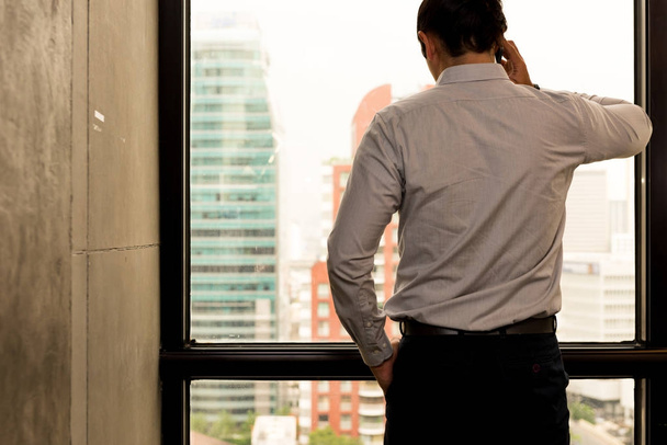 Young business leader concept. Young handsome business man in modern office on phone looking out the window. White male with stripe shirt with talking on mobile device. - 写真・画像