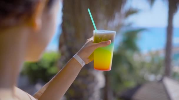 woman is holding glass with cocktail in background of resting area of a hotel near sea in sunny weather, making small sip - Video