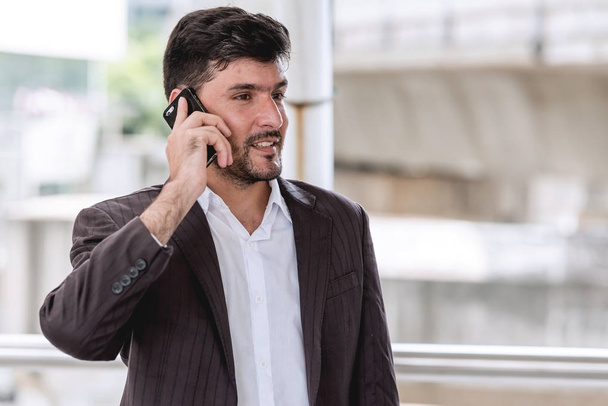 Mobile office concept.  Handsome successful white business man or sale man talking on his cell phone with financial building in background. Taken outdoor, apply urban looks effect. - Photo, Image
