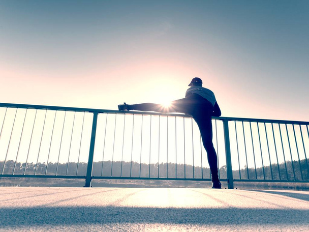 Morning runner in tall black leggings makes body stretching on bridge path. Outdoor exercising - Photo, Image