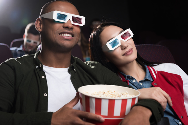 young interracial couple in 3d glasses with popcorn watching movie in cinema - Photo, image