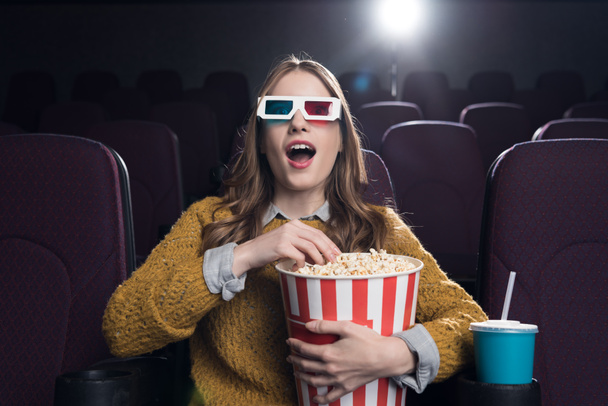 jonge opgewonden vrouw in 3d bril met grote mand van popcorn kijken naar film in de bioscoop - Foto, afbeelding