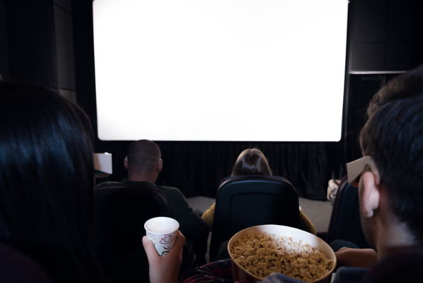 back view of friends with popcorn and drink sitting in cinema with white blank screen  - Foto, imagen