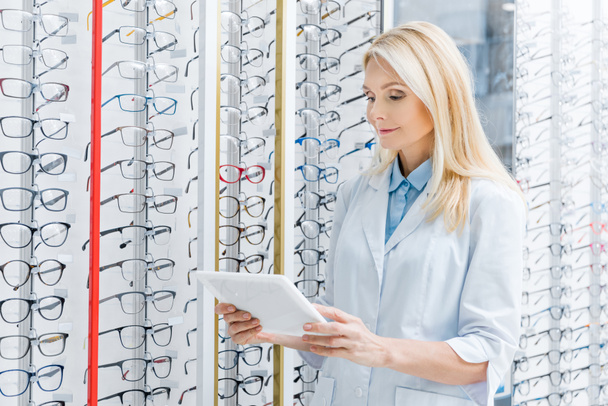 professional female ophthalmologist using digital tablet in optics with glasses on shelves - Foto, Imagem