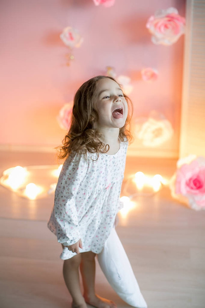 Little girl posing with facial expression holding white pillow with lights on background - Φωτογραφία, εικόνα