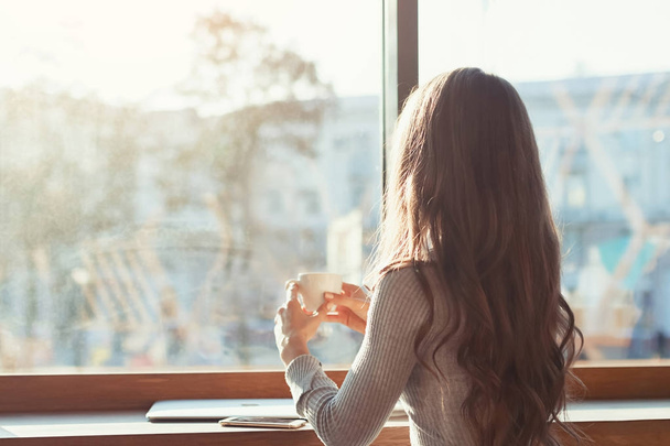 kafedeki kadına bir masada oturuyor, kahve içer ve pencerenin dışındaki şehir panoraması bakar. Bir güneşli sabah üzerinde bir kafede bir kız - Fotoğraf, Görsel
