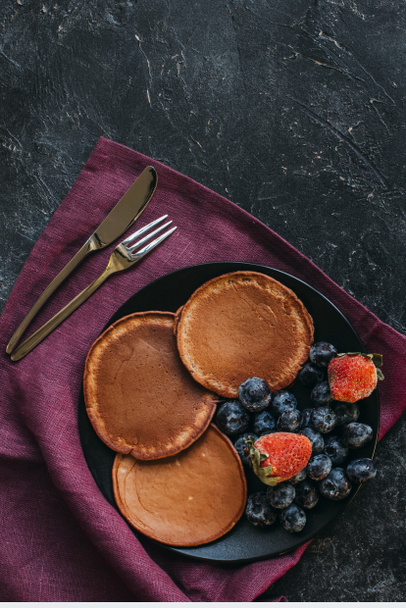 top view of chocolate pancakes with berries and cutlery on black concrete surface - Φωτογραφία, εικόνα