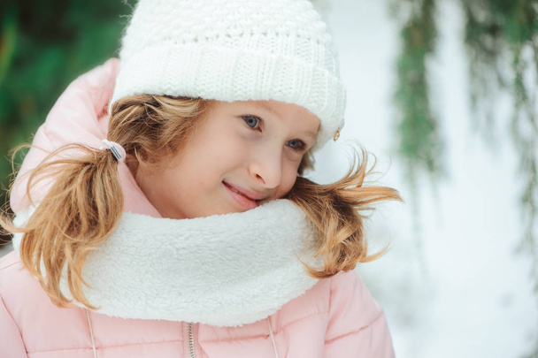 winter portrait of 8 years old kid girl walking outdoor in snowy day, wearing white knitted hat and pink coat - Foto, Bild