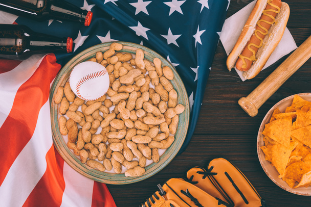 top view of baseball ball on plate with peanuts, baseball bat, glove, hot dog and beer bottles on american flag - Foto, imagen