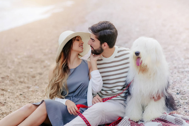 young couple in love resting with fluffy dog sitting on plaid on beach and smiling looking at each other - Foto, imagen