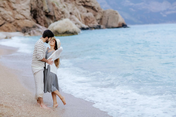 elegante giovane coppia innamorata che si abbraccia in piedi sulla spiaggia a piedi nudi con scogliera e montagna sullo sfondo, lunghezza intera
 - Foto, immagini