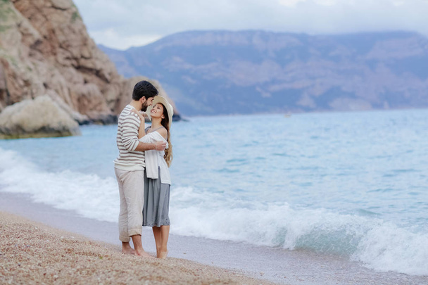 щасливий чоловік обіймає свою красиву усміхнену жінку, що стоїть на пляжі босоніж зі скелею і горою на фоні, повної довжини
 - Фото, зображення