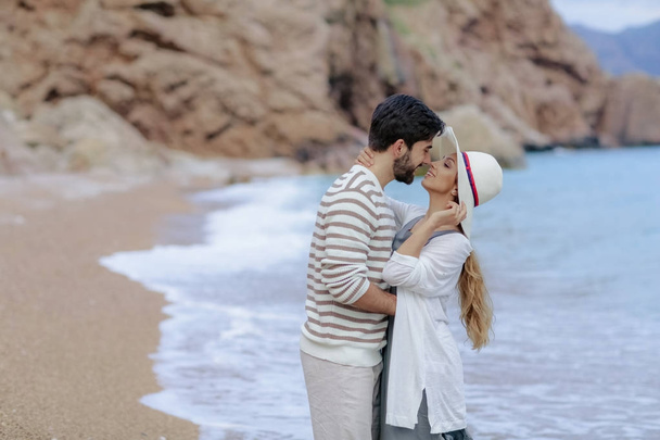 schöner Mann umarmt hübsches Mädchen mit weißem Hut, das zusammen am Meeresufer steht - Foto, Bild