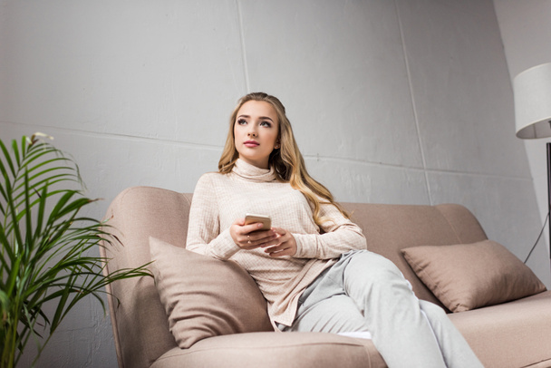 thoughtful young woman using smartphone on couch at home and looking away - Foto, imagen