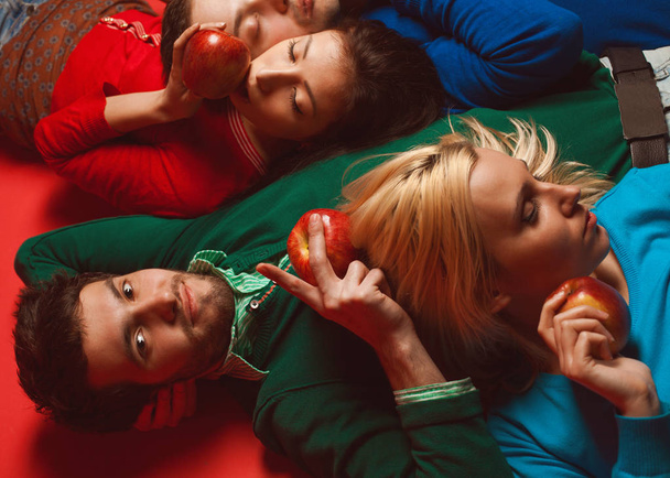 Picnic concept. Portrait of four stylish close friends hugging, having rest, holding red apples and lying on red background. Guys having fun. Hipster style. Studio shot - Photo, image