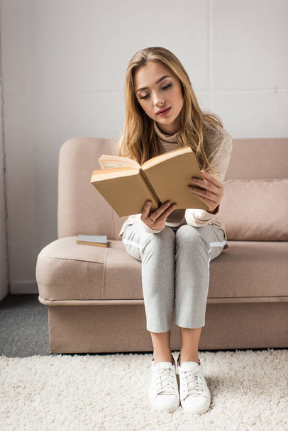 concentrated young woman reading book on cozy couch at home - Foto, Bild