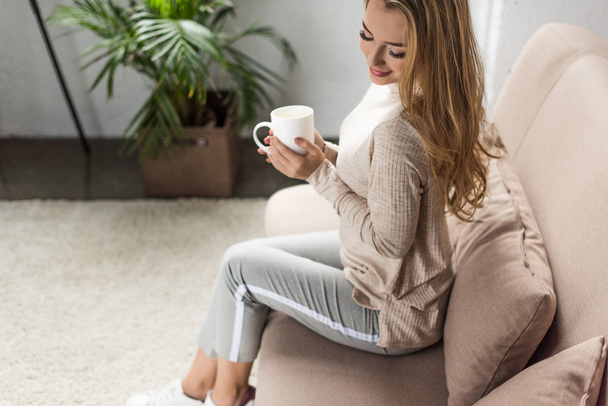beautiful young woman with cup of hot drink sitting on couch at home - Foto, Imagem