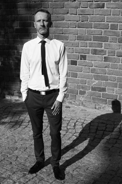 Smart casual handsome mature man stands over old house, building of European city and looking at camera. Hand in pockets. Graphic shadows. Monochrome outdoor shot - Foto, afbeelding