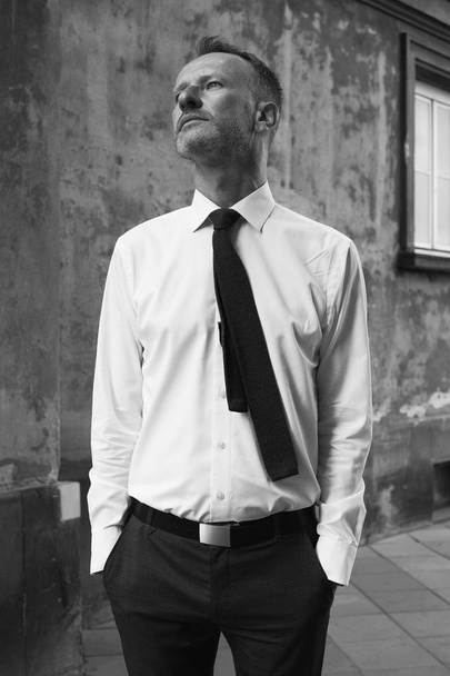 Smart casual handsome mature man stands over old house, building of European city and looking at camera. Hands in pockets. Monochrome outdoor shot - Valokuva, kuva