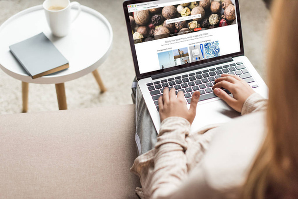 cropped shot of woman at home sitting on couch and using laptop with shutterstock website on screen - Fotografie, Obrázek