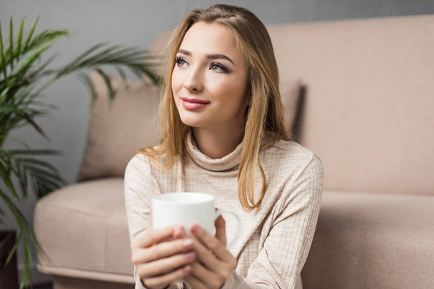 happy young woman in warm sweater with cup of hot drink at home and looking away - Photo, Image