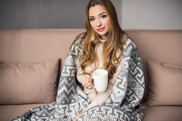 beautiful young woman covered in warm plaid drinking coffee on couch - Photo, Image