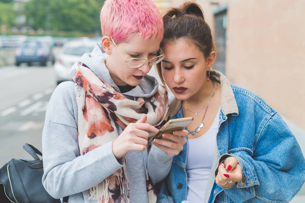 スマートフォンを使用して屋外で2人の若い女性-オンライン、若者文化、ソーシャルネットワークの概念 - 写真・画像