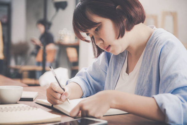 Młoda kobieta w białej sukni, siedząc przy stoliku w kawiarni i pisanie w notesie. Asian kobieta rozmawia smartphone i filiżankę kawy. Freelancer pracuje w kawiarni. Vintage efekt stylu Zdjęcia - Zdjęcie, obraz