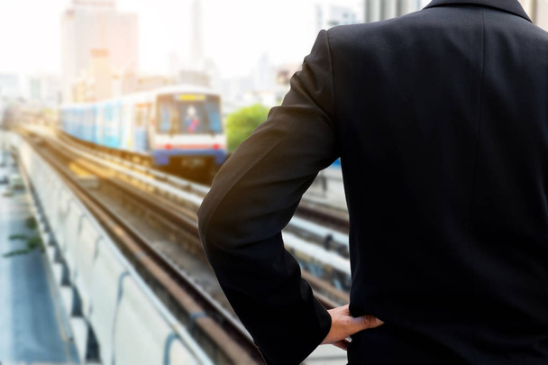 Hátoldalán üzletember állandó sötét szürke öltönyt és Csípőre tett kézzel várja a vonat, a vasútállomás a city városi csúcsforgalom. - Fotó, kép
