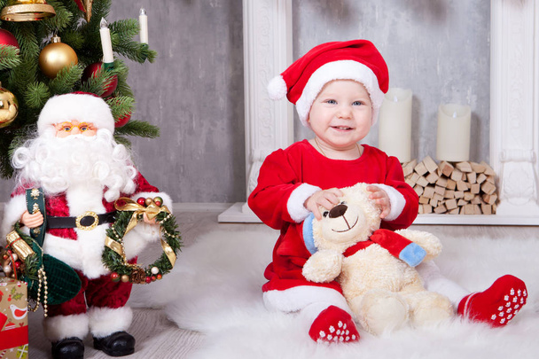 Christmas or New year celebration. Little girl in red dress and santa hat with bear toy sitting on the floor near the Christmas tree with xmas gifts. A fireplace with christmas stocking on background - Photo, Image