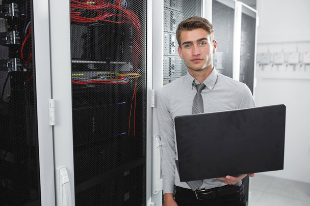 Retrato de un joven moderno sosteniendo la computadora portátil de pie en la sala de servidores trabajando con supercomputadora
 - Foto, imagen