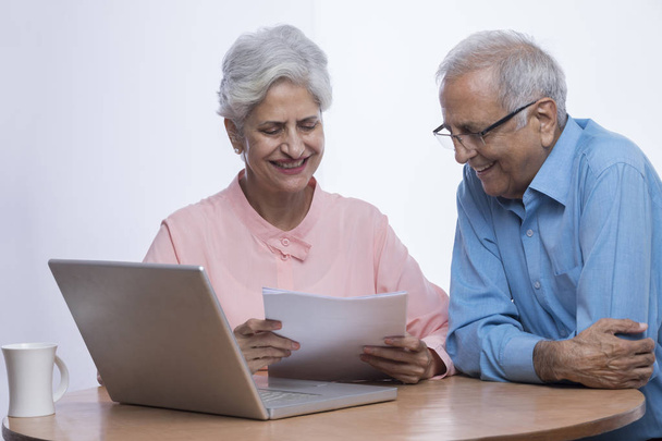 Felice coppia di anziani utilizzando il computer portatile e guardando i documenti seduti sul tavolo
 - Foto, immagini