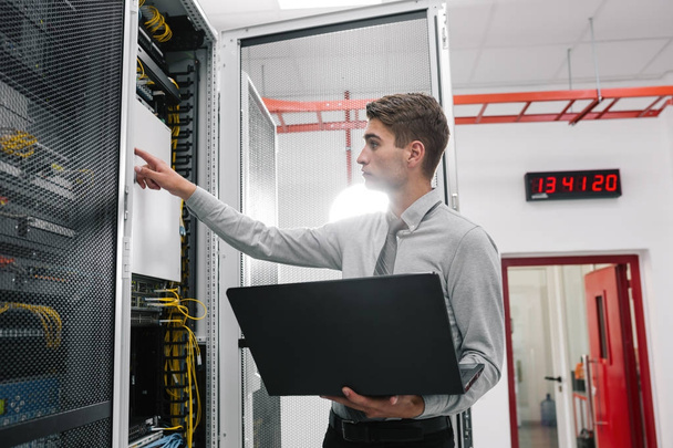 Portrait of modern young man holding laptop standing in server room working with supercomputer - Foto, Imagem