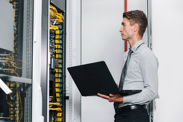 Ritratto di giovane uomo moderno che tiene il computer portatile in piedi nella stanza del server che lavora con il supercomputer
 - Foto, immagini