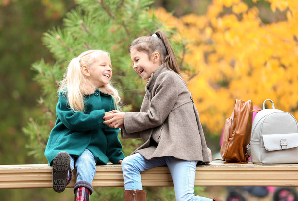 Cute little girls in outwear sitting and playing on bench outdoors - Zdjęcie, obraz