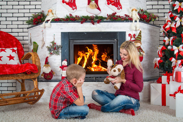 Merry Christmas and Happy Holidays!.Mother and son playing with a toy dog by the fireplace. - Фото, изображение