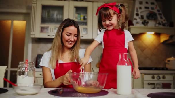 Mooie jonge moeder helpt haar dochtertje langs om te koken taart in rode schorten. Giet de bloem in een kom en klop het beslag om te maken van een taart in de keuken. - Video