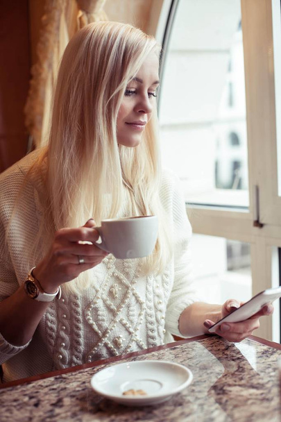 Genç güzel kadın bir fincan sıcak cappuccino tutuyor. Genç bir kadın kapuçino içiyor ve telefonda konuşurken rahat bir kafede oturuyor. Kız kakao içiyor. Kış. Rahat sonbahar. - Fotoğraf, Görsel