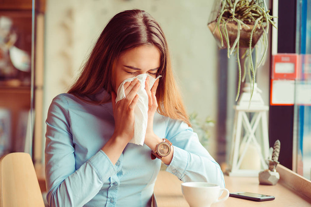 Grypy. Młoda kobieta dostał alergii nosa, kichanie nos grypy, siedzi przy stole w modna kawiarnia coffee shop filiżankę gorącego napoju, obok - Zdjęcie, obraz