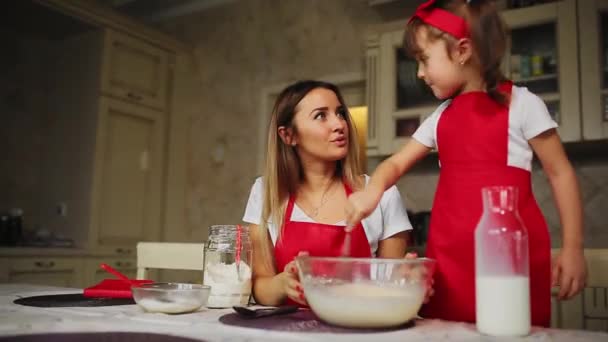 Krásná mladá matka pomáhá její malá dcera spolu se vařit dort v červené zástěry. Nasypte mouku do mísy a porazit těsto udělat dort v kuchyni. - Záběry, video