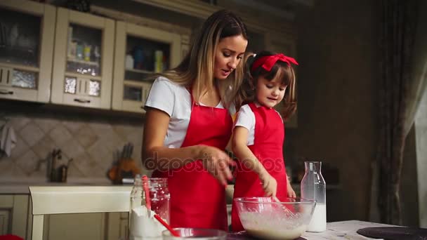 Die schöne junge Mutter hilft ihrer kleinen Tochter beim Kuchenbacken in roten Schürzen. Das Mehl in eine Schüssel geben und den Teig schlagen, um in der Küche einen Kuchen zu backen. - Filmmaterial, Video