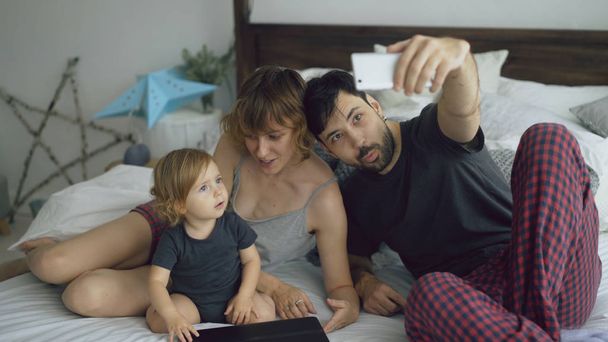 Young family with cute little girl taking selfie portrait on smartphone camera sitting in bed at home - Photo, Image