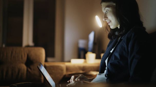 Mujer bonita joven usando el ordenador portátil y navegar por las redes sociales sentado en el entrenador en casa en la noche
 - Foto, Imagen