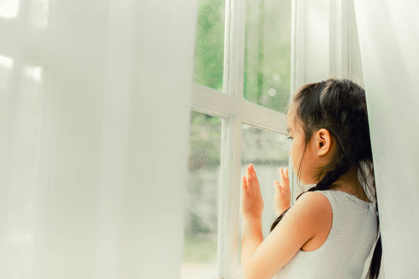 Criança triste, menina olhando a chuva caindo através de uma janela em casa
 - Foto, Imagem