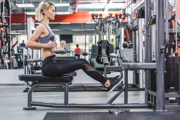 vista lateral de la mujer joven haciendo ejercicio con la máquina de fila en el gimnasio
 - Foto, Imagen