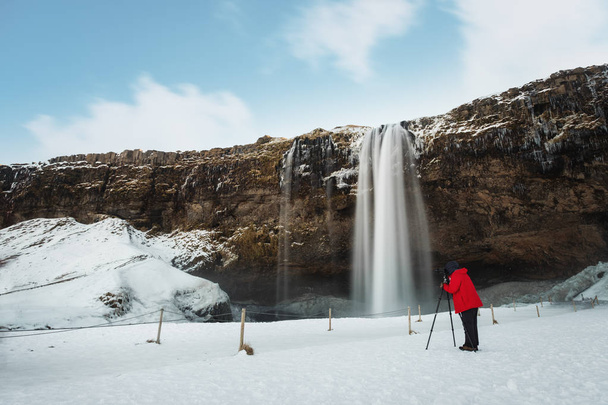 Winter waterfall landscape, photographer in red jacket taking photograph with camera tripod at Seljalandsfoss waterfall in Iceland - Photo, Image